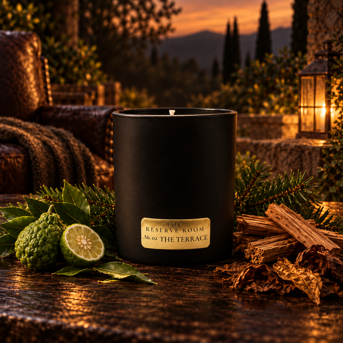 A matte black scented candle jar sits on a rustic wooden table outdoors at dusk, surrounded by green citrus, leaves, wood bark, and evergreen branches. The label reads 'RESERVE ROOM' and 'THE TERRACE'.