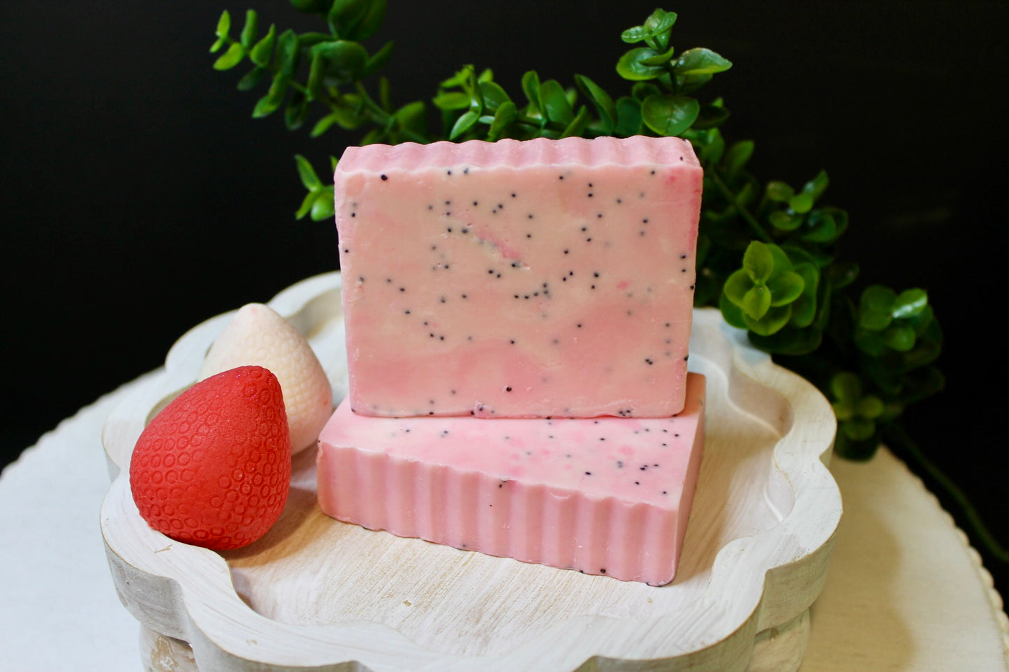 Two pink soap bars with specks on a white stand with a strawberry and green leaves in the background.