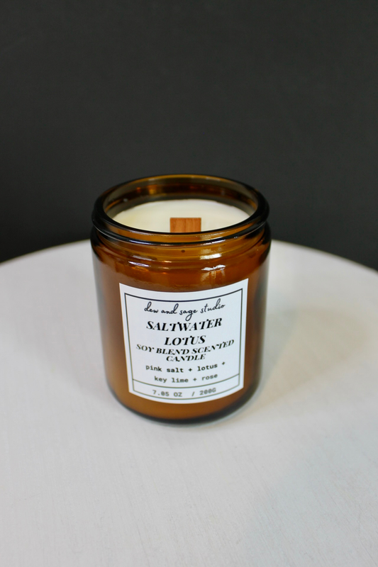 An amber glass jar candle with a white label reading 'dew and sage studio' and 'SALTWATER Lotus SOY BLEND SCENTED CANDLE' on a white surface against a dark gray background.