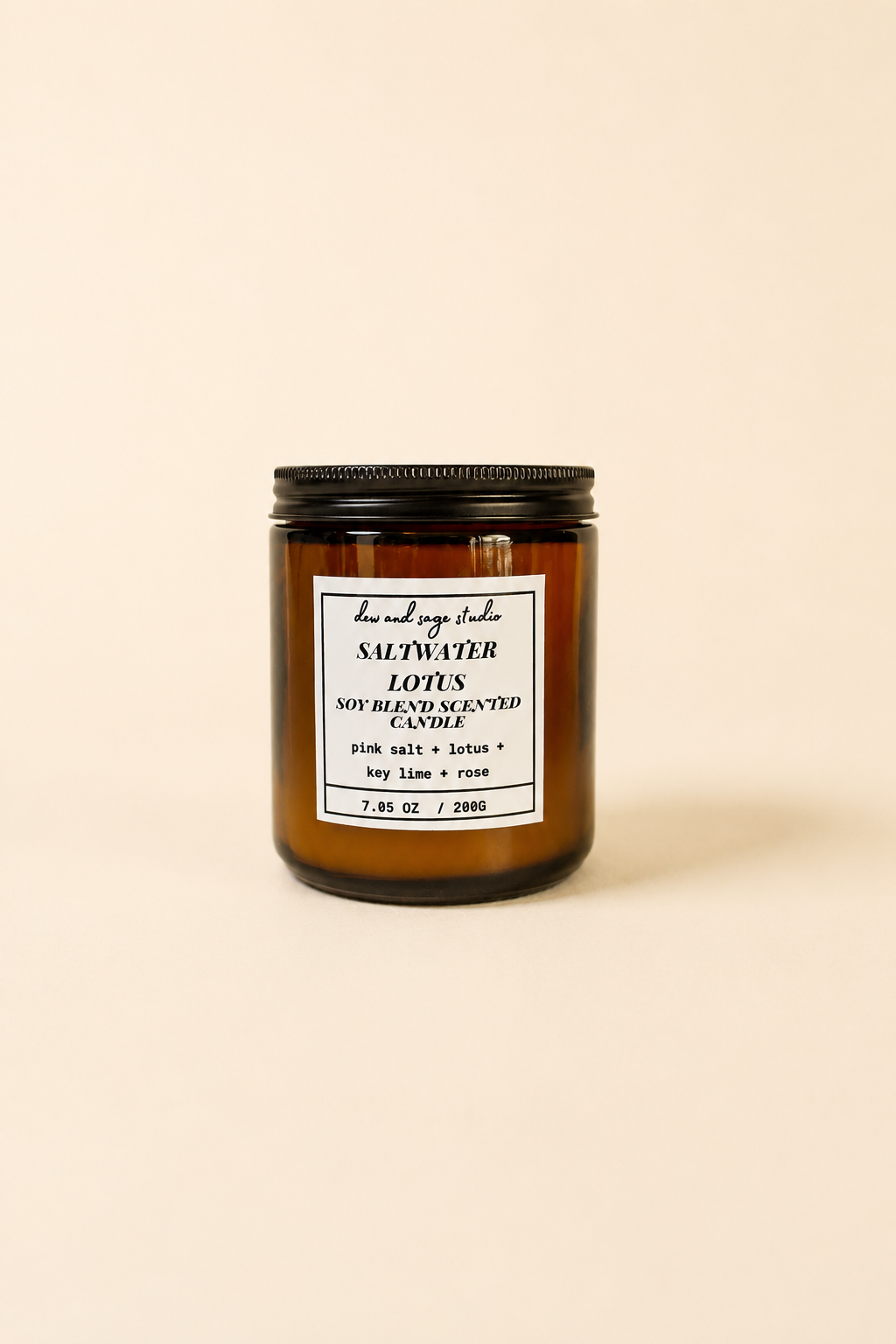 An amber glass jar candle with a black lid and a white label reading 'SALTWATER' and 'LOTUS', photographed on a plain warm beige background.