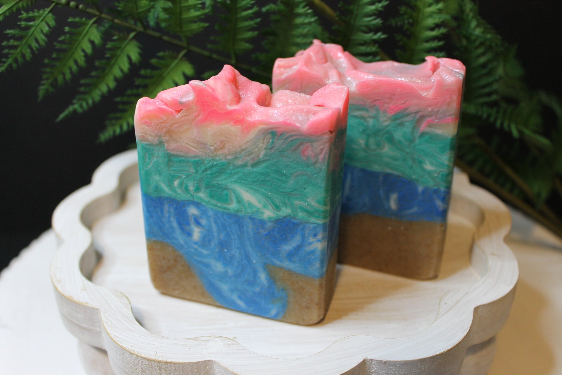 Two ocean layered soap bars with pink, blue, and green colors on a white stand with a green leafy background.
