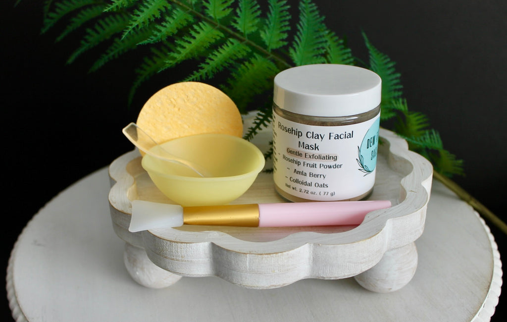 Cosmetic products including a jar, brush, and mask on a decorative stand with ferns in the background.