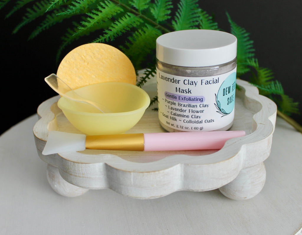 Facial mask jar with a sponge, brush, and facial mask on a wooden stand with a fern in the background.