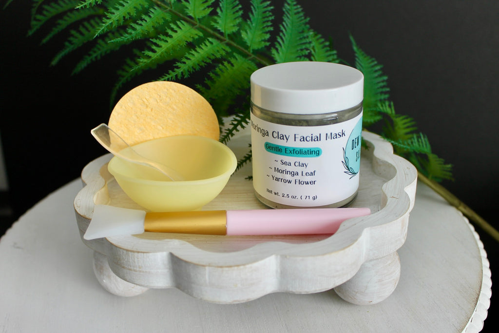 Facial mask products including a jar, sponge, and mask on a decorative stand with ferns in the background.