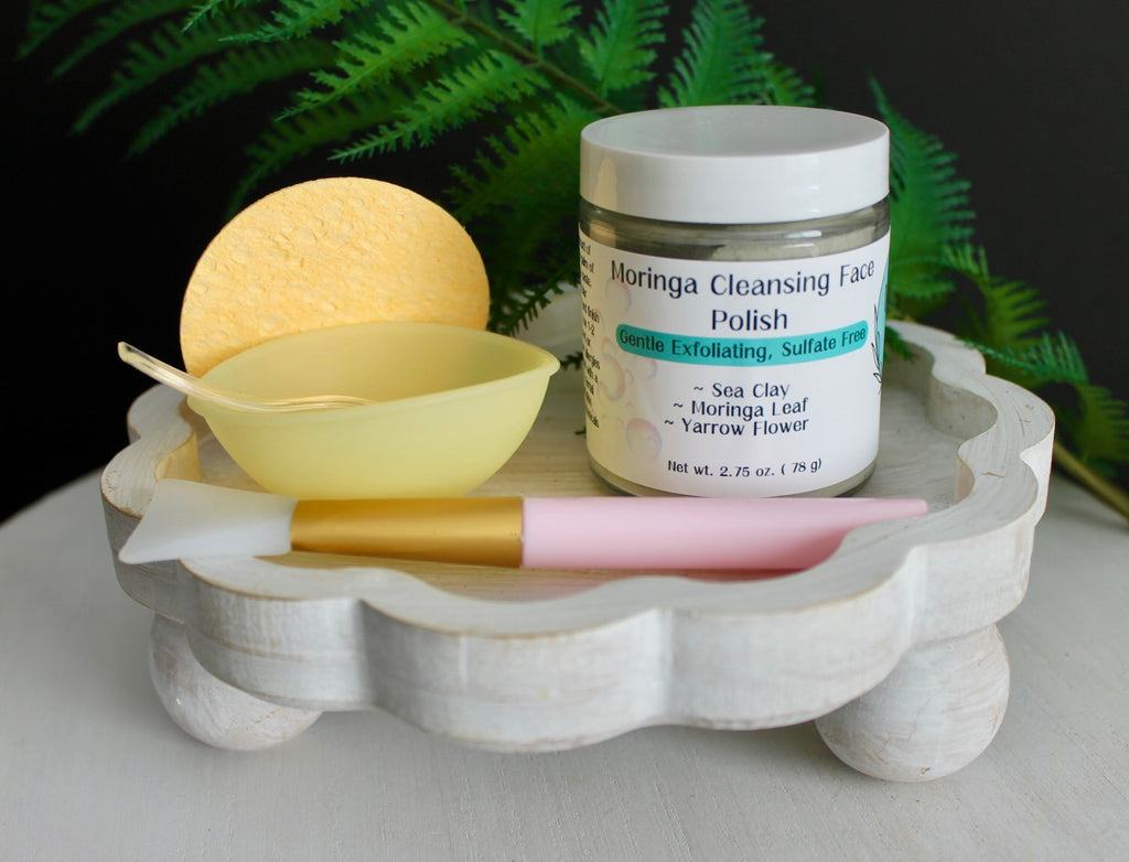 Cosmetic products including a jar of 'Moringa Cleansing Face Polish', a yellow ball, a bowl, and a pink brush on a white stand with a green leafy background.