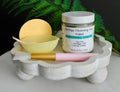 Cosmetic products including a jar of 'Moringa Cleansing Face Polish', a yellow ball, a bowl, and a pink brush on a white stand with a green leafy background.