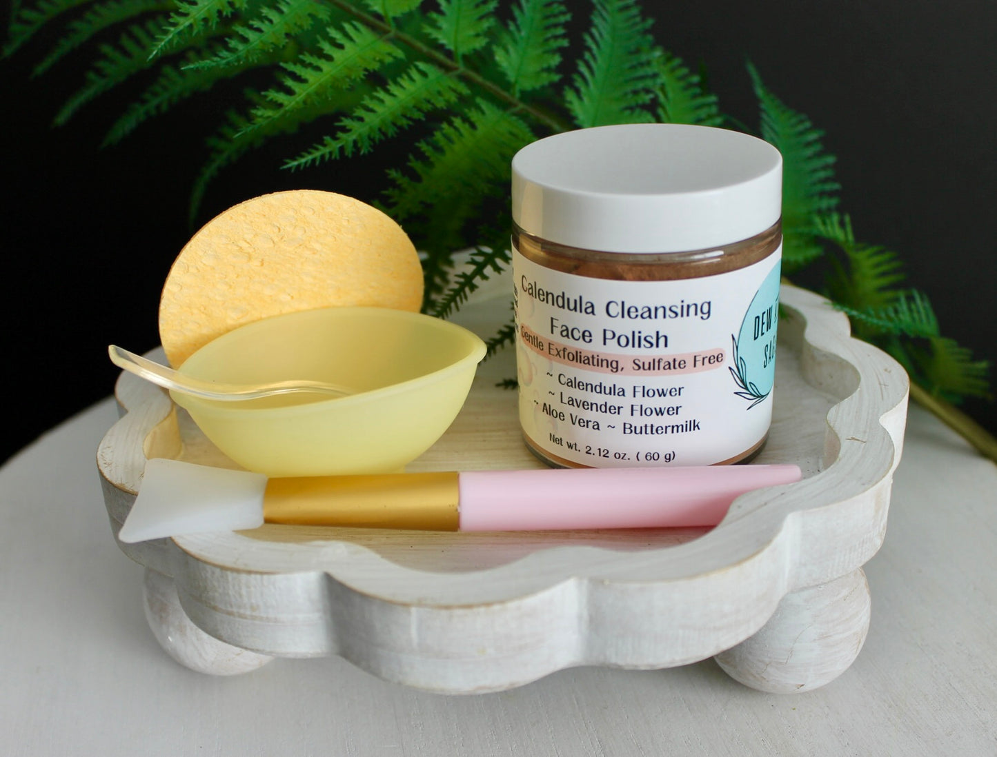 Cosmetic products including a jar, brush, and sponge on a decorative tray with ferns in the background.
