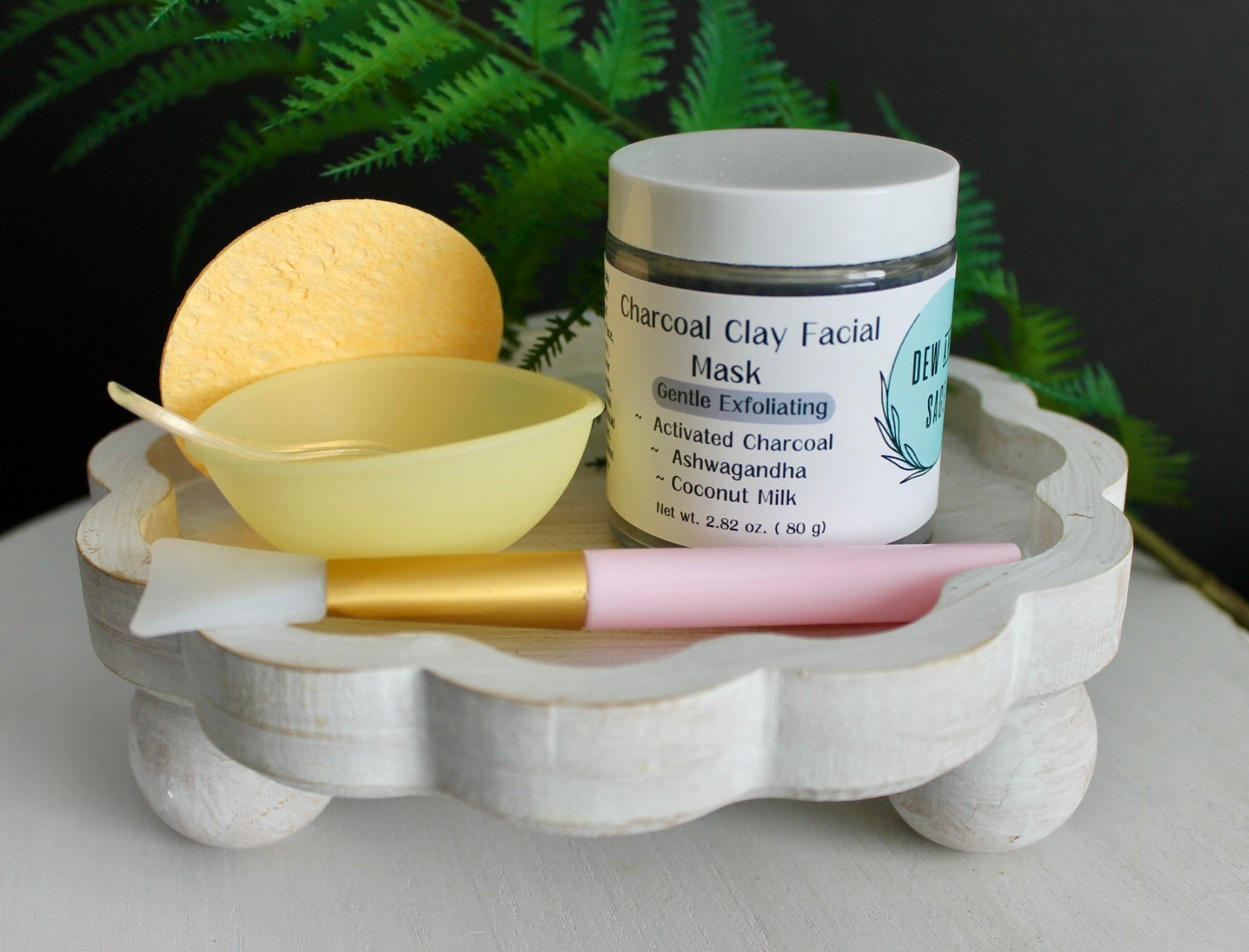Cosmetic items including a jar of charcoal clay facial mask, a bowl, sponge and an applicator on a decorative stand with a fern in the background.