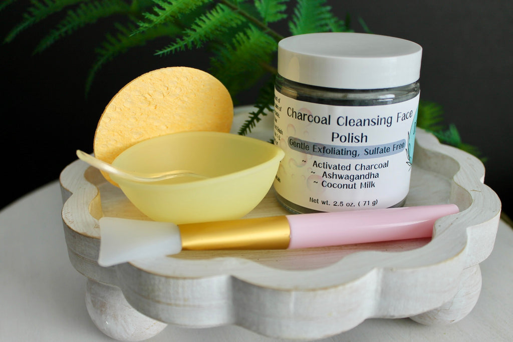 Cosmetic products including a jar of 'Charcoal Cleansing Face Polish', a yellow sponge, and a pink brush on a decorative stand with a green leafy background.
