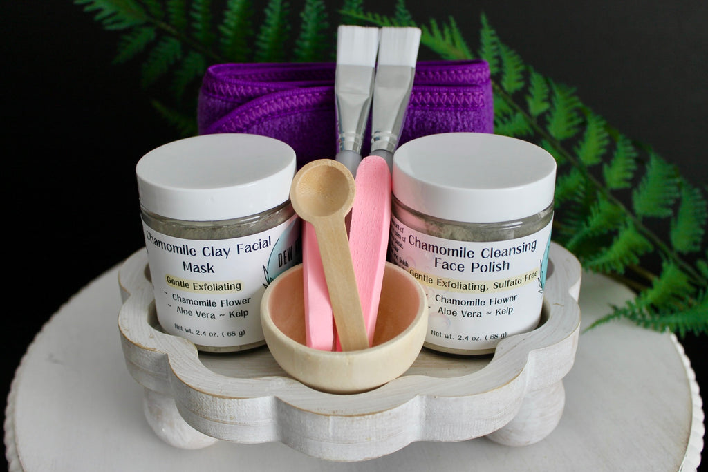 Cosmetic set with jars labeled 'Chamomile Clay Facial Mask' and 'Chamomile Cleansing Face Polish' on a white stand.