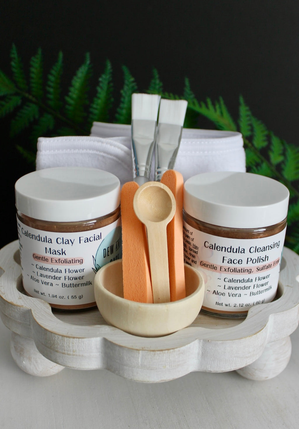 Skincare products including jars, a brush, and a towel on a wooden tray with ferns in the background.