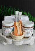Skincare products including jars, a brush, and a towel on a wooden tray with ferns in the background.