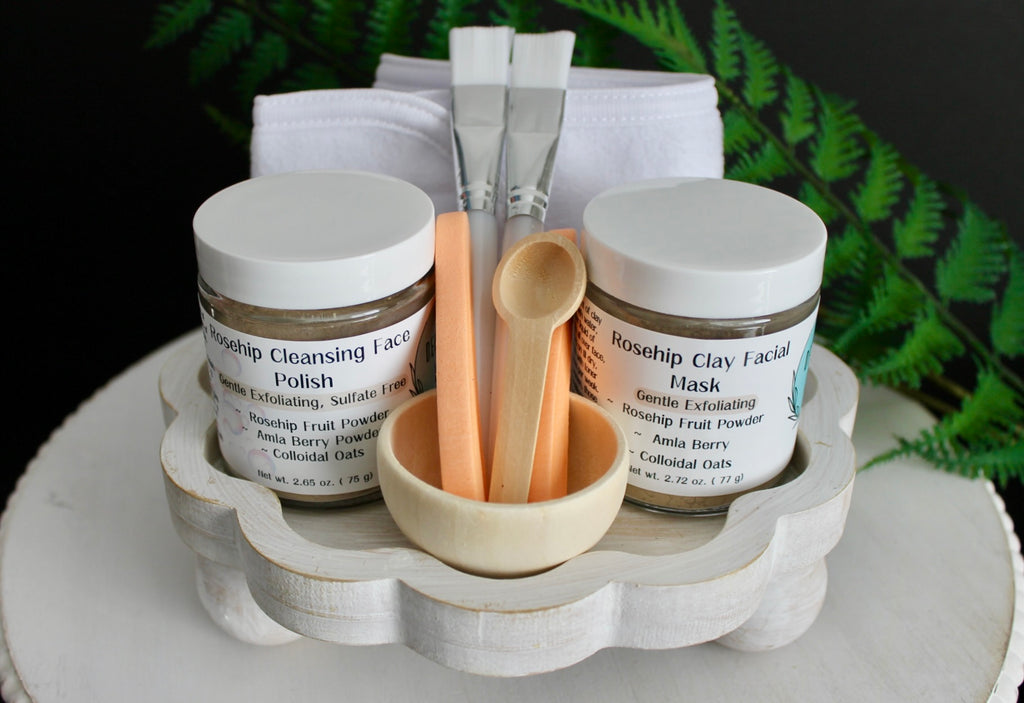 Cosmetic set with jars, brushes, and a wooden bowl on a white stand with greenery in the background.