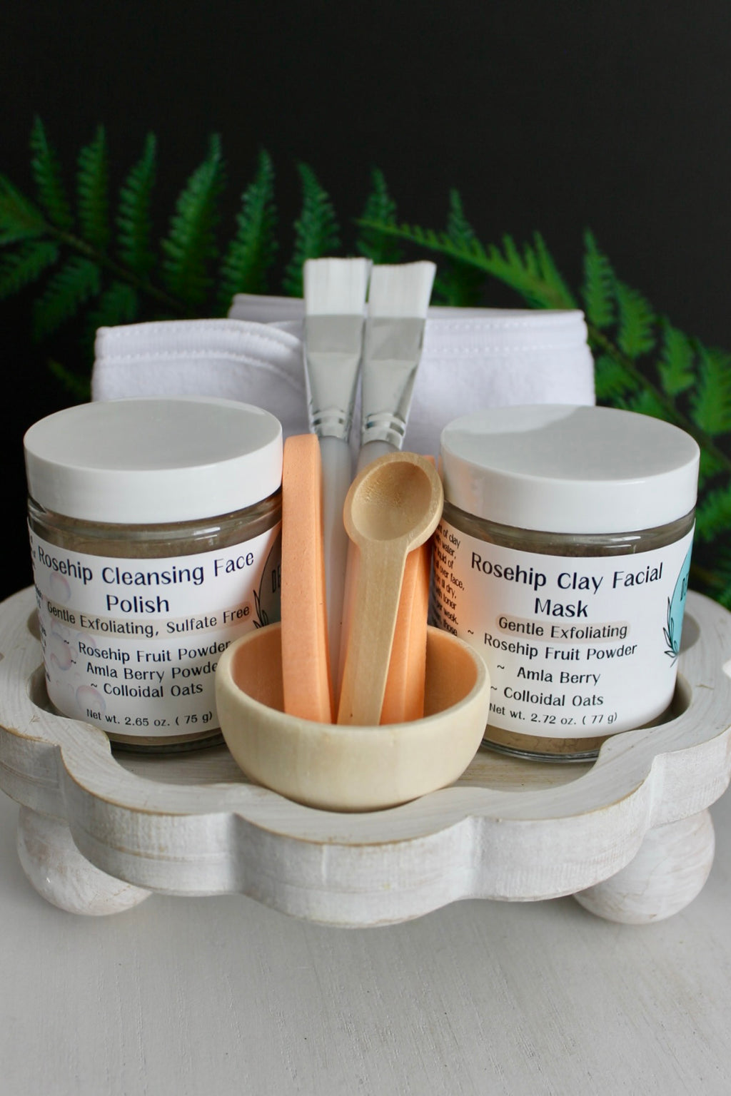 Cosmetic set with jars, brushes, and a bowl on a wooden stand with greenery in the background.