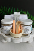 Cosmetic set with jars, brushes, and a bowl on a wooden stand with greenery in the background.