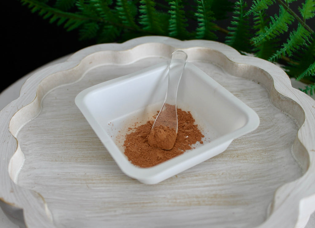 Small white dish with brown powder and a spoon on a textured surface with greenery in the background