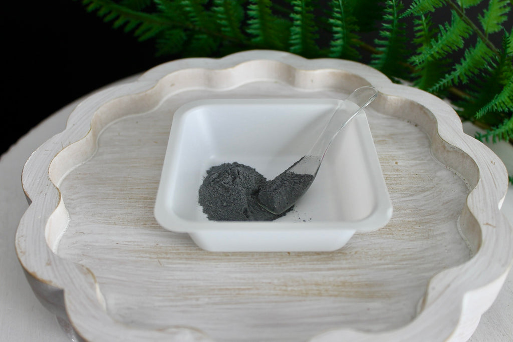 White ceramic dish with a small amount of dark powder inside, placed on a textured white surface with greenery in the background.