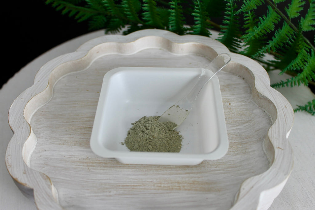 White ceramic bowl with green powder on a white surface with ferns in the background