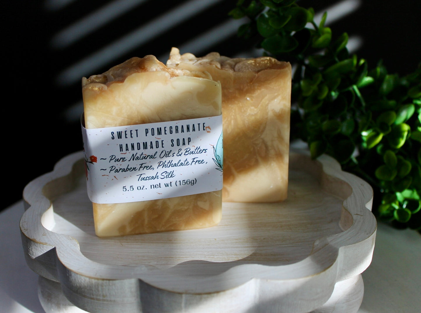 Two handmade soap bars on a white stand with a dark background and greenery.