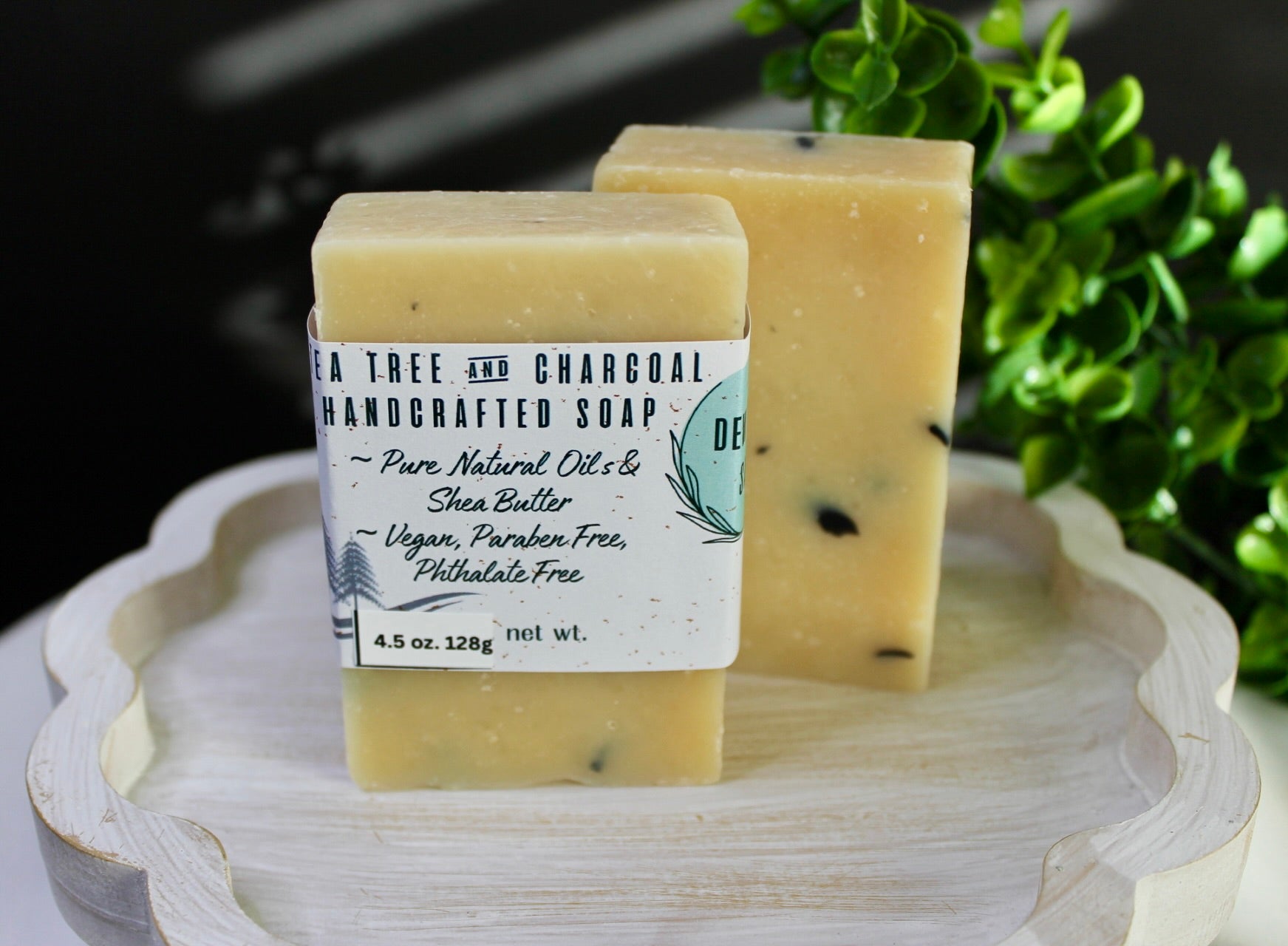 Two bars of handmade soap on a wooden tray with a plant in the background
