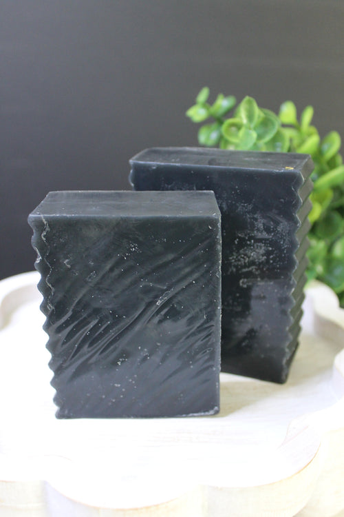 Two bars of dark soap stacked on a white surface with a blurred green plant in the background