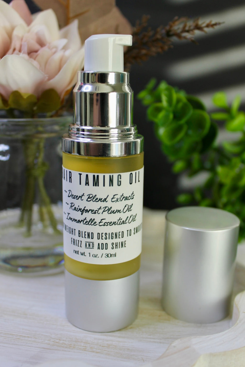 Skincare bottle labeled 'Hair Taming Oil' on a wooden surface with flowers and greenery in the background.