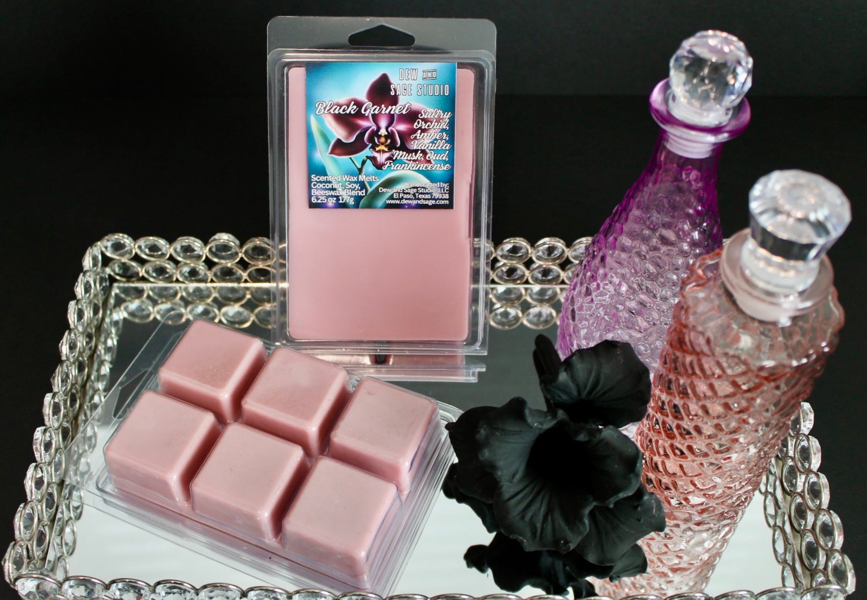 Pink wax melts with a decorative tray, black flowers, and a purple perfume bottle on a dark background.