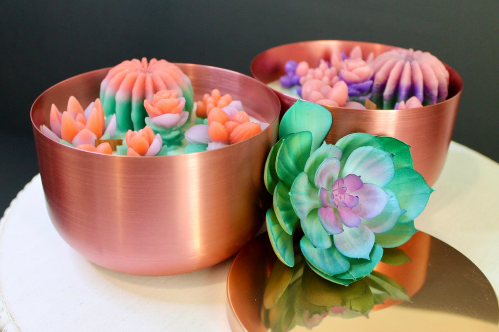 Decorative flower-shaped candles in copper bowls on a reflective surface with a dark background.