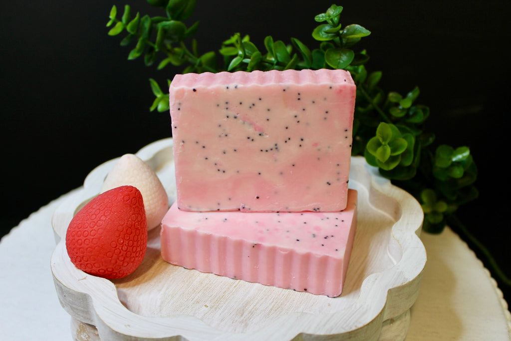 Two pink soap bars with specks on a white stand with a strawberry and green leaves in the background.