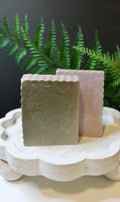 Two green and purple soap bars on a decorative stand with a fern in the background