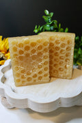 Two honeycomb-patterned soap bars on a white stand with greenery in the background