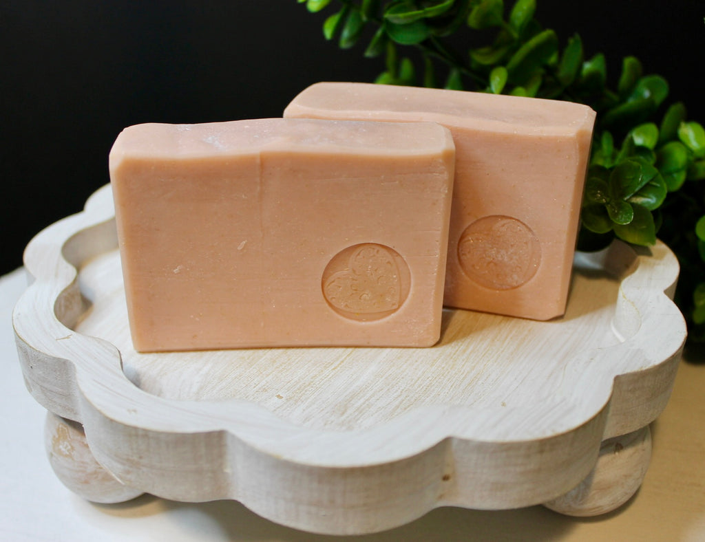 Two pinky beige bars of soap on a decorative white stand with a blurred green plant in the background.