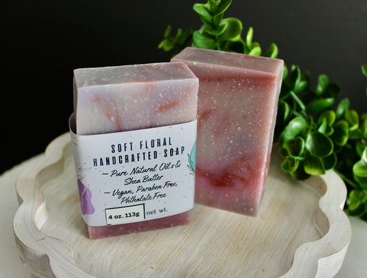 Two bars of soft floral handcrafted soap on a wooden stand with greenery in the background.