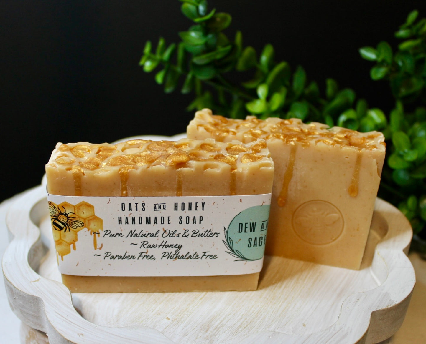 Two bars of handmade soap with labels on a wooden stand against a dark background with greenery.