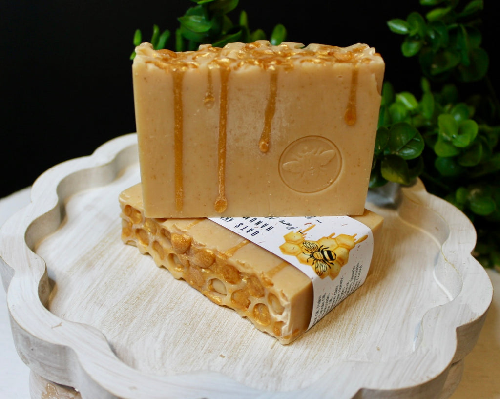 Two bars of honey-scented soap on a decorative white plate with a blurred green plant background.