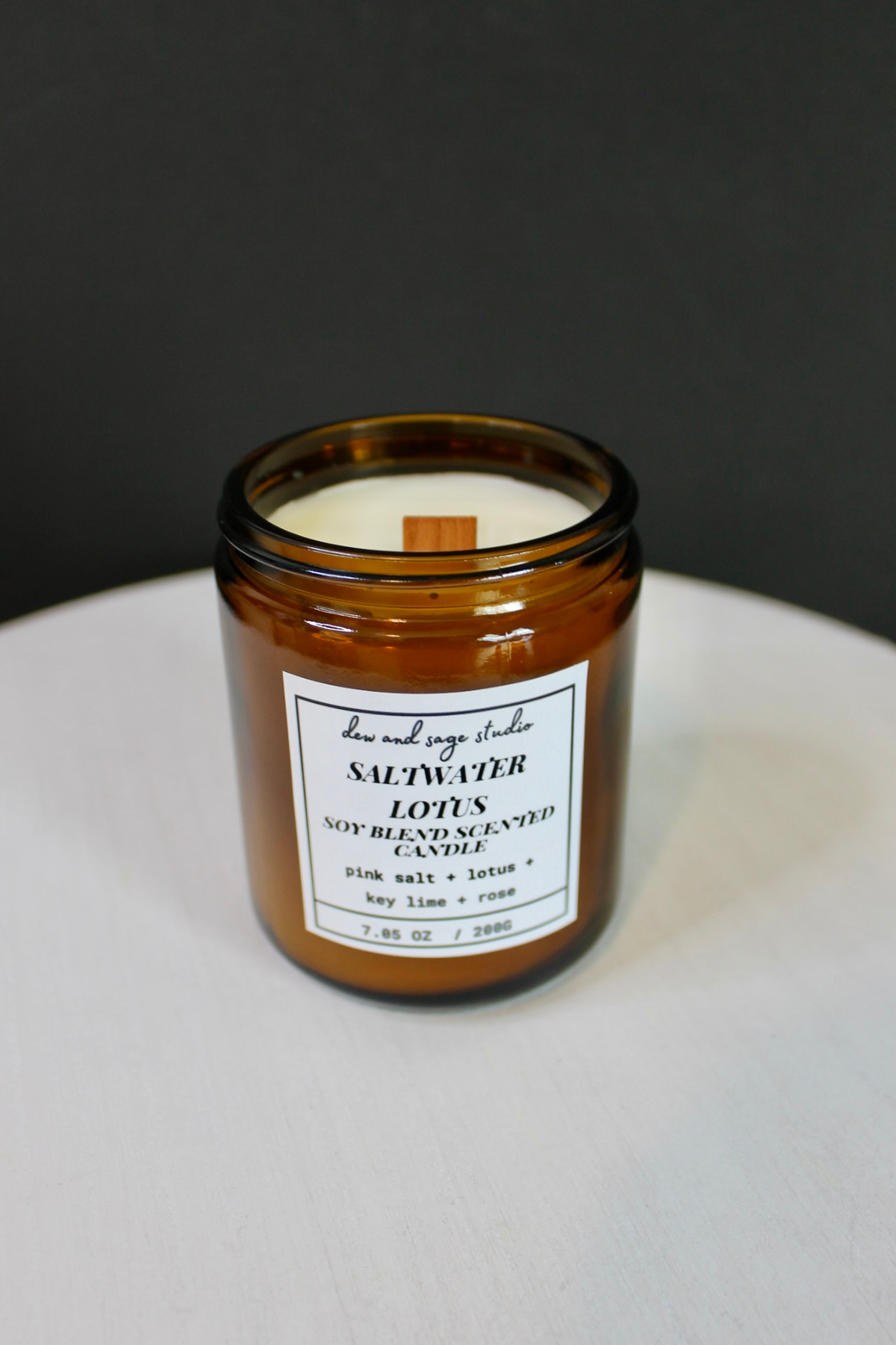 An amber glass jar candle with a white label reading 'dew and sage studio' and 'SALTWATER Lotus  SOY  BLEND SCENTED  CANDLE' on a white surface against a dark gray background.