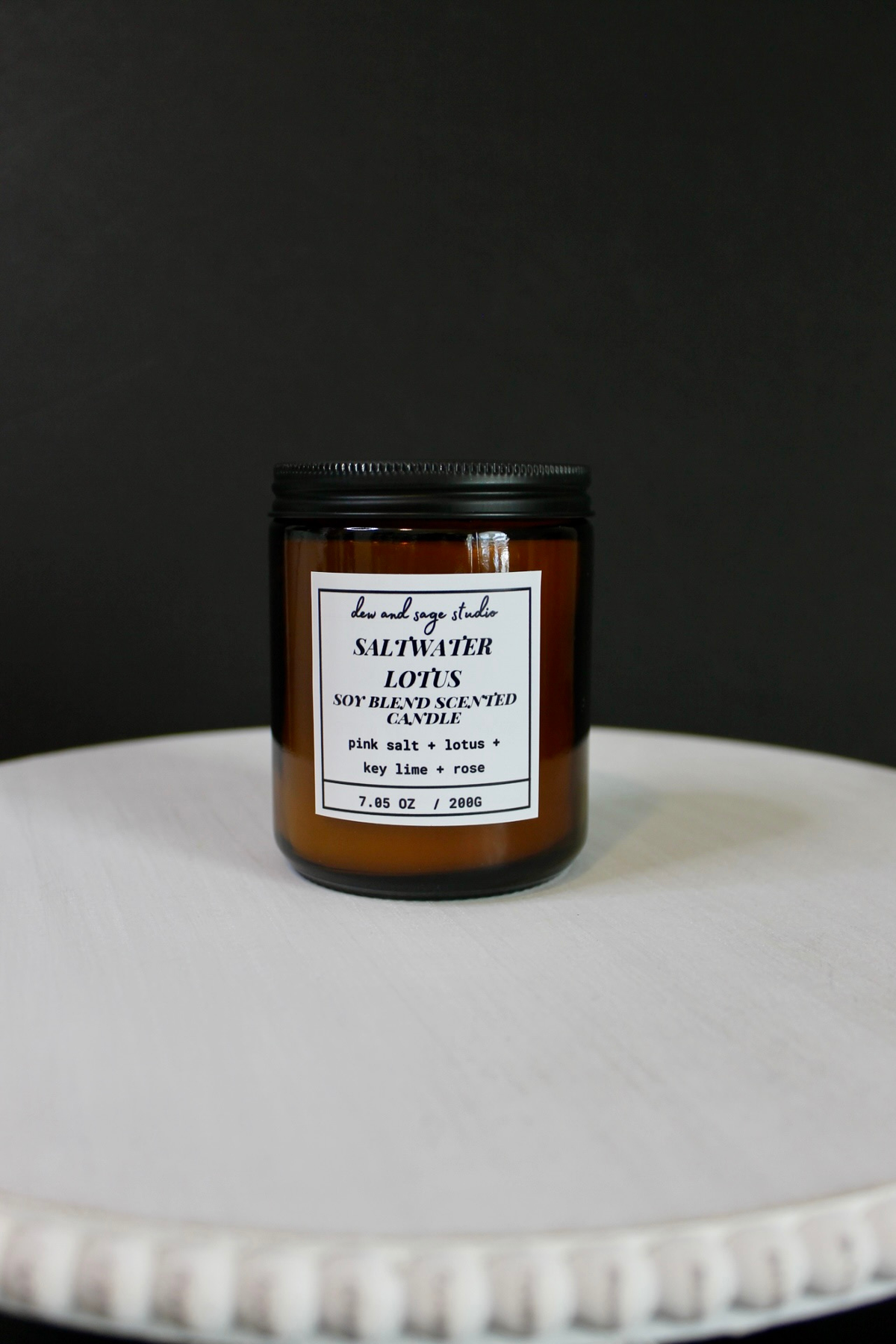 An amber glass jar candle with a black lid and a white label reading 'SALTWATER LOTUS' by 'the real sage studio', placed on a white pedestal against a dark background.